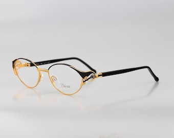 May include: Oval-shaped spectacles featuring a gold-tone metal frame with black accents on the outer corners and temples. The right lens displays the word Diva in a black script font. The temples are thin and black, connecting to the gold frame.