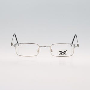 Pode incluir: Óculos retangulares prateados com armações de metal finas e pontas das hastes pretas. A lente direita exibe um pequeno adesivo preto com o texto "X-eyes" impresso em branco. A ponte e as dobradiças têm um padrão texturizado sutil.