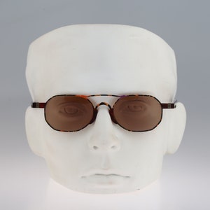 Puede incluir: Un par de gafas de sol de carey marrón con lentes marrones. Las gafas tienen un marco de metal y están en una cabeza de maniquí blanca.