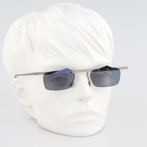 May include: Rimless sunglasses featuring a thin metallic brow bar and slender temples. The lenses are rectangular and tinted dark gray. Attached to the top of the frame are two small, geometric decorative accents, one in purple and one in black.