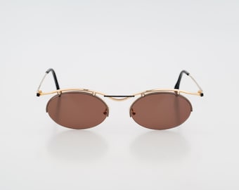 Puede incluir: Gafas de sol ovaladas con lentes marrones y montura dorada. Las patillas son finas y negras. Las gafas de sol tienen un estilo retro y vintage.