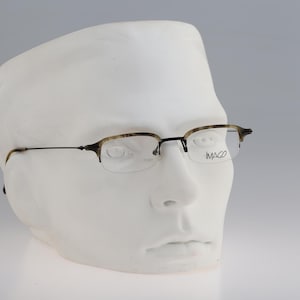 Puede incluir: Un par de gafas de media montura marrón y negro con lentes transparentes. Las gafas están en una cabeza de maniquí blanca. El nombre de la marca "IMAGO" está impreso en la lente derecha.