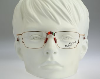 May include: Gold-tone metal eyeglasses with a rectangular frame, featuring red nose pads and a bridge with red and green accents. The right lens displays the black text Robert Rudger. The temples have small red accents at the tips.