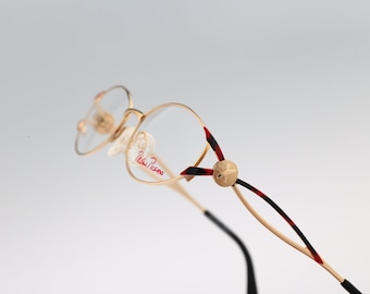 May include: Gold-tone metal spectacles featuring round frames with the text Paloma Picasso printed in red on the lens. The temples have a decorative gold sphere accent and a black and red tortoiseshell pattern along the thin metal arms.