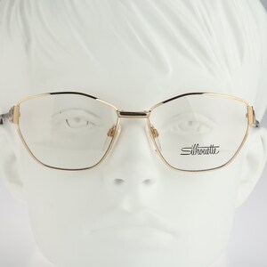 Könnte beinhalten: Goldfarbene Metallbrille mit Cat-Eye-Gestell und klaren Gläsern. Der Markenname "Silhouette" ist auf dem rechten Glas aufgedruckt.