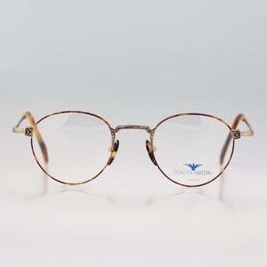 Può includere: Un paio di occhiali rotondi in tartaruga con montatura in metallo argentato. Il nome del marchio "Dakota Smith" è stampato sulla lente destra.
