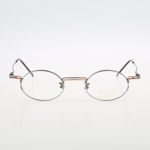 May include: Oval-shaped eyeglasses with a thin, silver-colored frame and a gold-colored bridge. The temples have a unique, decorative design. The lenses are clear, and the text "TAXI MADE IN JAPAN" is visible on the right lens.
