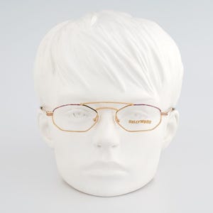 Puede incluir: Gafas doradas con doble puente y montura geométrica. Las lentes están delineadas en dorado, con detalles morados en la parte superior de la montura. La marca "Hollywood" está impresa en la lente derecha. Las gafas se exhiben en una cabeza de maniquí blanca.