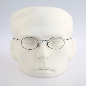 May include: Oval-shaped eyeglasses with a thin, dark frame are displayed on a white mannequin head. The glasses have a delicate design, with the lenses held in place by a slender frame. The overall aesthetic is minimalist and classic.