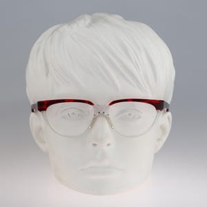 Puede incluir: Gafas con montura tipo browline que presenta un patrón de carey en rojo oscuro y negro en los bordes superiores y lentes transparentes. La parte inferior de las lentes no tiene montura y se sujeta mediante un puente de metal plateado.