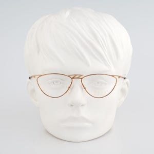 May include: Gold-toned eyeglasses with a cat-eye shape, displayed on a white mannequin head. The frames have a delicate, thin design. The glasses are set against a plain white background, highlighting their elegant style.