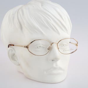 May include: Gold-tone metal eyeglasses with a geometric frame shape. The temples feature a small CD logo near the hinges, and the right lens is etched with the text Christian Dior. The earpieces are finished in a brown tortoiseshell pattern.