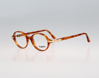 Puede incluir: Gafas con patrón de carey, monturas redondas y detalles de metal en tono dorado en las varillas. El cristal derecho presenta el nombre de la marca Byblos impreso en texto negro. Estas gafas tienen varillas finas y un puente clásico.