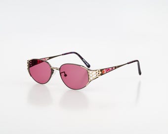 Puede incluir: Gafas de sol con lentes de color rosa oscuro y monturas de metal dorado. Las patillas presentan un diseño decorativo de celosía cerca de las bisagras y una incrustación con patrón floral en los laterales. Las puntas son marrón oscuro.