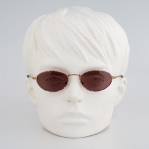 May include: Oval-shaped sunglasses with a gold-colored frame and dark red lenses. The lenses are set in a decorative, patterned rim. The sunglasses are displayed on a white mannequin head.