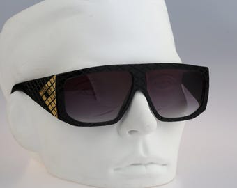May include: Black sunglasses with a wide, flat-top frame featuring a textured, grid-like pattern across the front and temples. The temples include a gold-colored, metallic, diamond-patterned accent near the hinges. The lenses are dark and gradient.