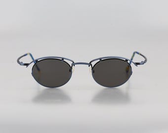 May include: A pair of round sunglasses with a matte blue metal frame. The design features a double-bridge structure where the upper bar arches over the circular dark gray lenses. The thin temples extend outward from the frame, showing a blue finish.