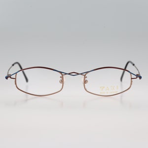 May include: A pair of rimless glasses with a brown and blue metal frame. The glasses have a subtle curve to the frame and say "TAXI" on the right lens.