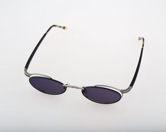 May include: Oval-shaped sunglasses featuring dark tinted lenses and a thin silver-tone metal frame. The temples are black with a tortoiseshell pattern at the tips. The bridge connects the two oval lenses, and the frame has a distinct layered design.