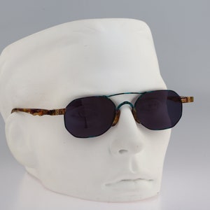 Puede incluir: Un par de gafas de sol grises oscuras con un marco estampado en marrón y verde. Las gafas de sol están en una cabeza de maniquí blanca.