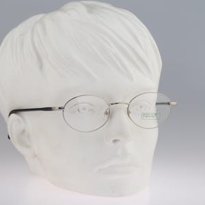 Puede incluir: Gafas de metal plateado con monturas ovaladas y patillas finas. El cristal derecho presenta una pequeña pegatina rectangular con el texto United Colors of Benetton. Las gafas se muestran sobre una cabeza de maniquí blanca y sin rasgos.
