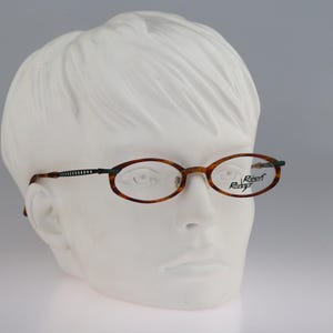 May include: Oval-shaped eyeglasses featuring a brown tortoiseshell pattern on the frames and thin, dark metal temples with a textured, ribbed design. The right lens displays the brand name Robert Rudger printed in a black, stylized script font.