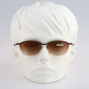 May include: Brown-rimmed sunglasses with a gradient lens tint, featuring a thin metal frame and a small rectangular sticker on the right lens with the text Calvin Klein. The eyewear is displayed on a white, featureless mannequin head.