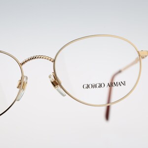 May include: Gold-rimmed round eyeglasses with a twisted bridge and Giorgio Armani branding on the right temple.