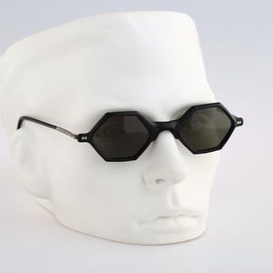 May include: Black octagon-shaped sunglasses with dark lenses on a white mannequin head.