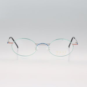 May include: Silver rimless oval eyeglasses with a blue and red accent on the arms. The brand name "T.A.S.C.I." is printed on the right lens.