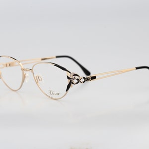 Op de afbeelding: Gouden metalen bril met zwarte accenten en een delicaat ontwerp. De monturen hebben een cat-eye vorm en de merknaam "Diva" is gedrukt op de rechter poot.