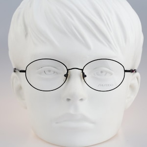 May include: Oval-shaped eyeglasses with a thin black metal frame. The clear lenses feature the Shiseido brand name printed in small black letters on the right lens. The glasses are displayed on a white mannequin head with a neutral facial expression.