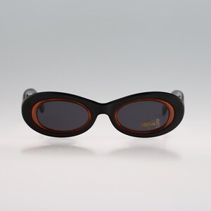May include: Black oval sunglasses with a brown rim and dark lenses. The sunglasses have the word "VERSACE" printed in gold on the right lens.