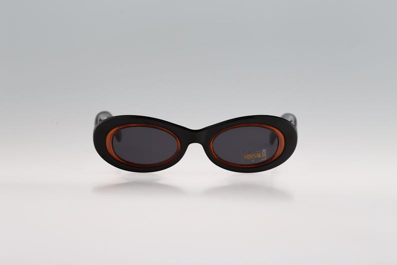May include: Black oval sunglasses with a brown rim and dark lenses. The sunglasses have the word "VERSACE" printed in gold on the right lens.