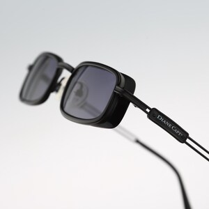 May include: Black sunglasses with rectangular frames and dark tinted lenses. The temples feature a black rectangular accent piece with the text DIANE CAPT printed in white. The design includes thin metal arms and adjustable nose pads.