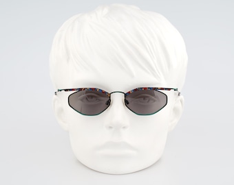 Puede incluir: Gafas de sol geométricas con un tinte gris oscuro. Las monturas son negras con un ribete verde y patillas estampadas de colores. Las gafas de sol se muestran en una cabeza de maniquí blanca.