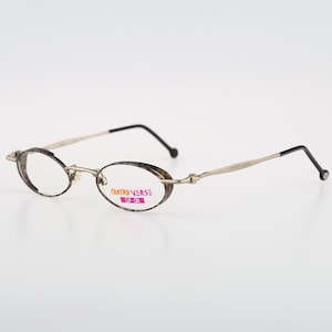 Puede incluir: Gafas ovaladas de color plateado con una patilla negra. Las gafas tienen lentes transparentes y la marca "OUTRO VERSE" está impresa en la montura.