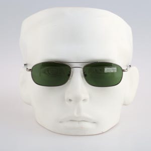 May include: Rectangular sunglasses with a thin silver metal frame and dark green tinted lenses. The right lens features a small rectangular sticker with the text Calvin Klein printed in black. The glasses are displayed on a white mannequin head.
