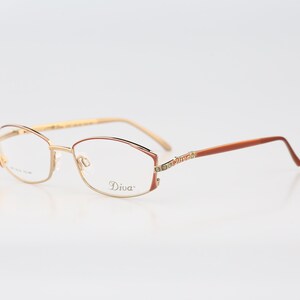 May include: Gold-toned metal eyeglasses with oval-shaped frames. The temples feature a brown finish and a decorative section with small embedded crystals. The right lens is printed with the word Diva in a cursive script font in black ink.