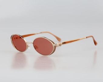 May include: Oval sunglasses featuring a thin gold-tone metal frame and reddish-brown tinted lenses. The temples are slender with a matching gold-tone finish and integrated hinges, providing a lightweight structure for these classic eyewear frames.