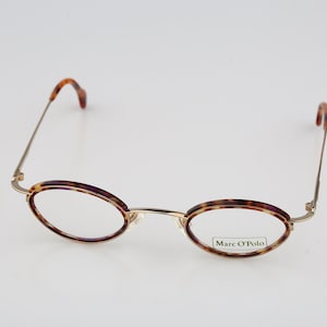 May include: Oval-shaped eyeglasses with a thin gold-tone metal frame and a brown tortoiseshell pattern on the rims. The right lens features the text Marc O'Polo printed in black. The temples are thin gold metal with tortoiseshell tips.