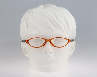 Puede incluir: Gafas de forma ovalada con una montura de color ámbar translúcido y patillas oscuras. Las lentes son transparentes y la palabra MaxMara está impresa en letras pequeñas y oscuras en la parte inferior de la lente derecha. Las gafas están sobre una cabeza blanca.
