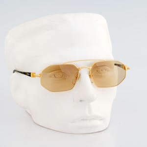 Puede incluir: Gafas de sol con montura dorada, doble puente y lentes octogonales. Las lentes son de color marrón claro y las patillas tienen un diseño negro y dorado. Las gafas se exhiben en una cabeza de maniquí blanca.
