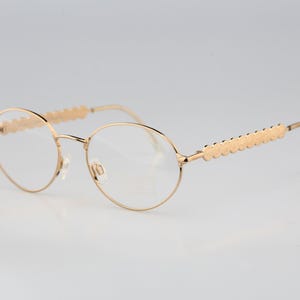 May include: Gold-toned metal eyeglasses featuring round lenses and thin frames. The temples are designed with a repeating scalloped or circular pattern along the outer edges, leading to curved earpieces. The bridge is thin and arched.
