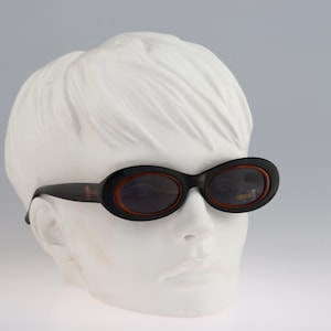 May include: A pair of black oval sunglasses with brown frames. The sunglasses are on a white mannequin head.