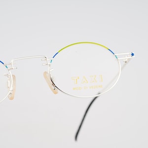 May include: Silver-toned metal eyeglasses with oval-shaped lenses. The upper rims feature a thin lime green and blue accent. The right lens is printed with the gold text TAXI MODI DI VEDERE. The frames include beige nose pads and black temple tips.