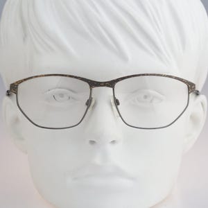 Könnte beinhalten: Brille mit dunkelbraunem Rahmen, der am oberen Rand ein gesprenkeltes, strukturiertes Muster aufweist, sowie dünnen Metallrändern unten. Die Gläser sind klar und transparent und werden durch einen Steg und Nasenpads auf einem weißen Kopf gehalten.