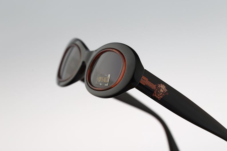 May include: Black oval sunglasses with a brown rim and a small logo on the side. The logo is a stylized image of a lion's head.