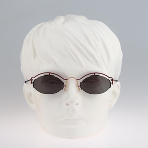 May include: A pair of oval-shaped sunglasses with a burgundy frame and dark gray lenses. The sunglasses are on a white mannequin head.
