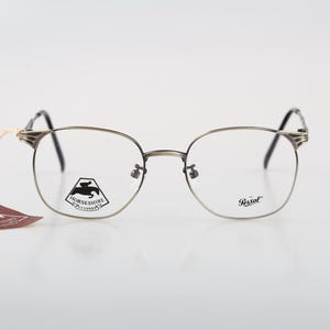 Puede incluir: Gafas de metal plateado con montura fina y puntas de patilla oscuras. El cristal derecho presenta un gráfico negro de un caballo y un jinete con el texto HORSESHIRE, mientras que el izquierdo muestra el logotipo de Persol. Lleva una etiqueta marrón.